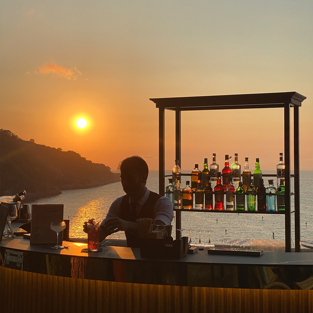 Barman al bancone bar con bottigliera dietro al tramonto